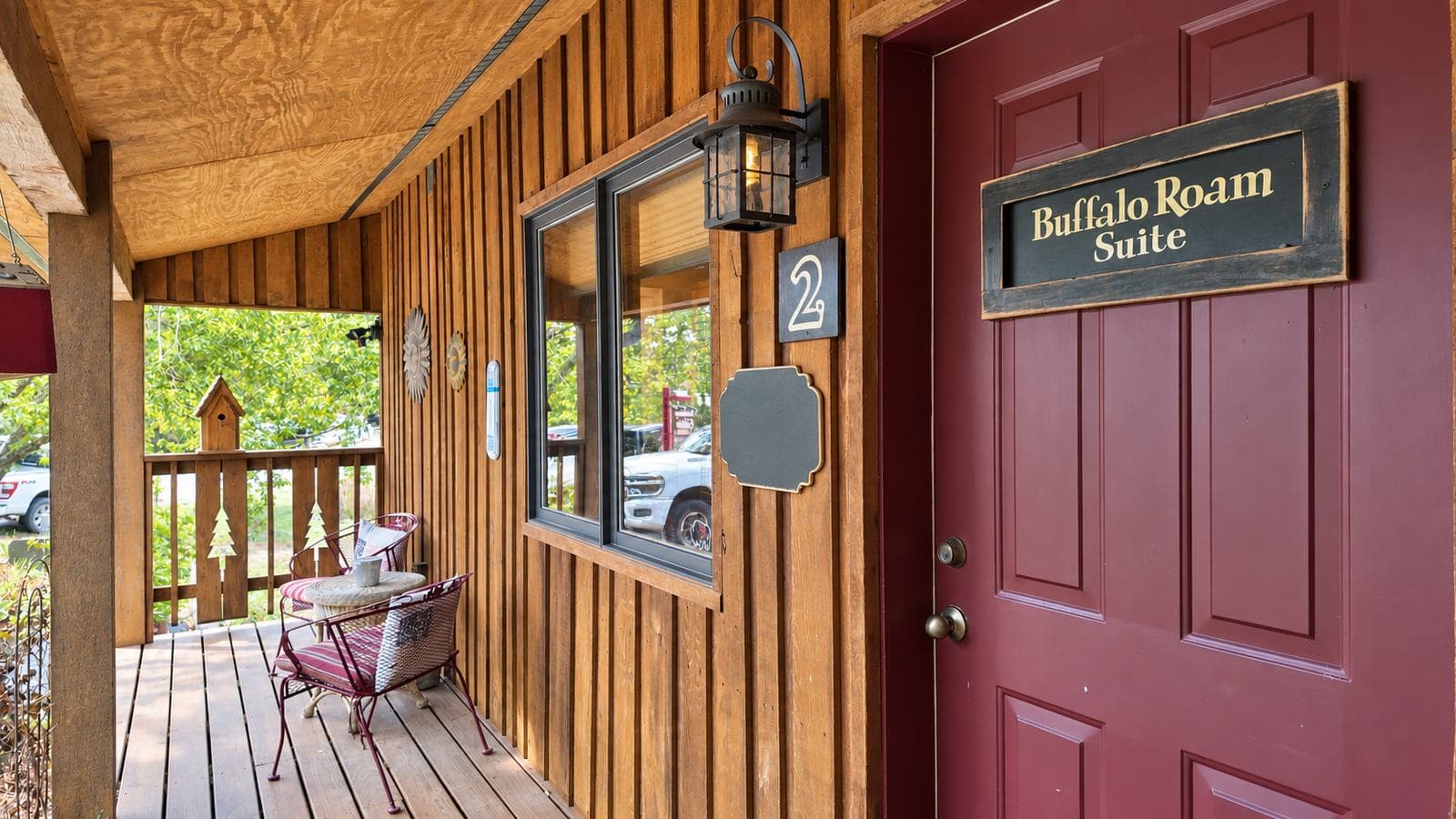 A wooden porch with a red door labeled "Buffalo Roam Suite" and two chairs set against a rustic background.