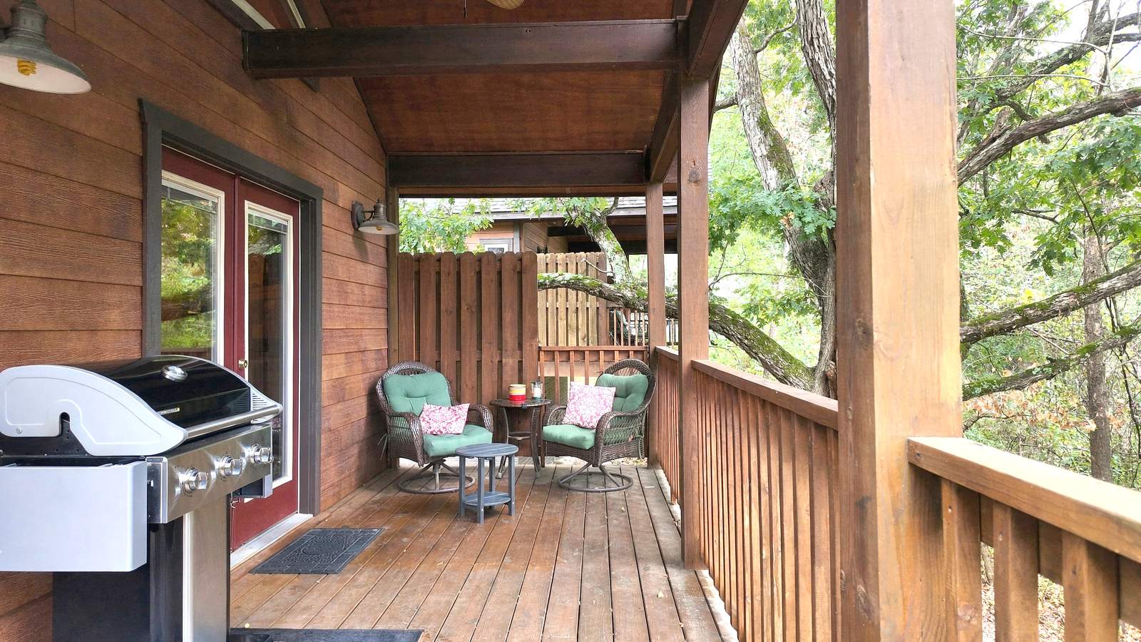 A cozy wooden deck features a grill, a small table, and two green chairs surrounded by trees.