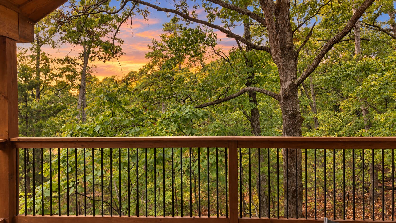 A wooden balcony overlooks a lush forest with vibrant trees and a colorful sunset sky.