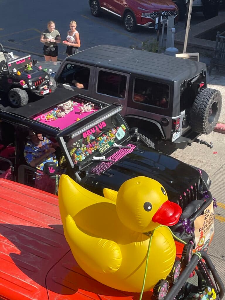 A large inflatable yellow duck sits on a decorated vehicle, with two women nearby and other cars in the background.