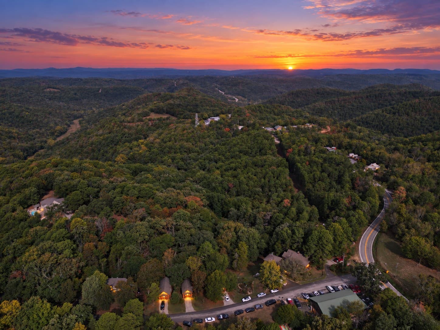 Aerial view of rolling hills and a winding road at sunset, with lush green trees and a colorful sky.