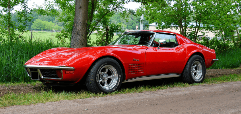 Red vintage sports car parked beside a gravel road surrounded by trees.