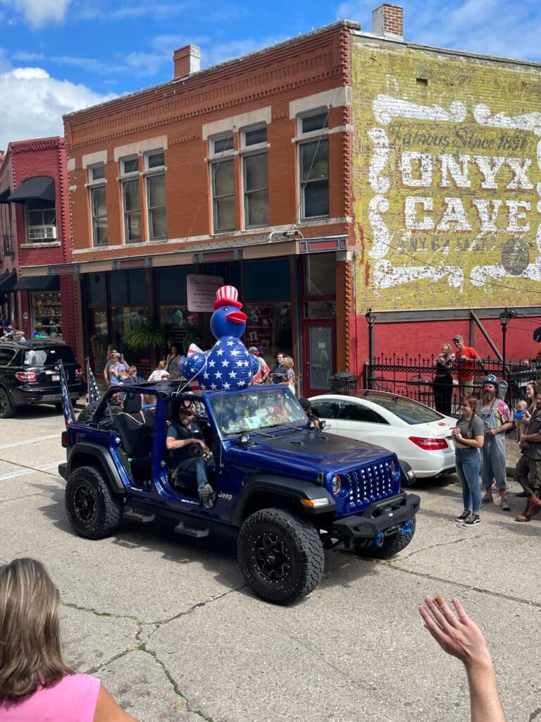 A blue Jeep with a large, patriotic rubber duck on top drives through a crowd at a parade.