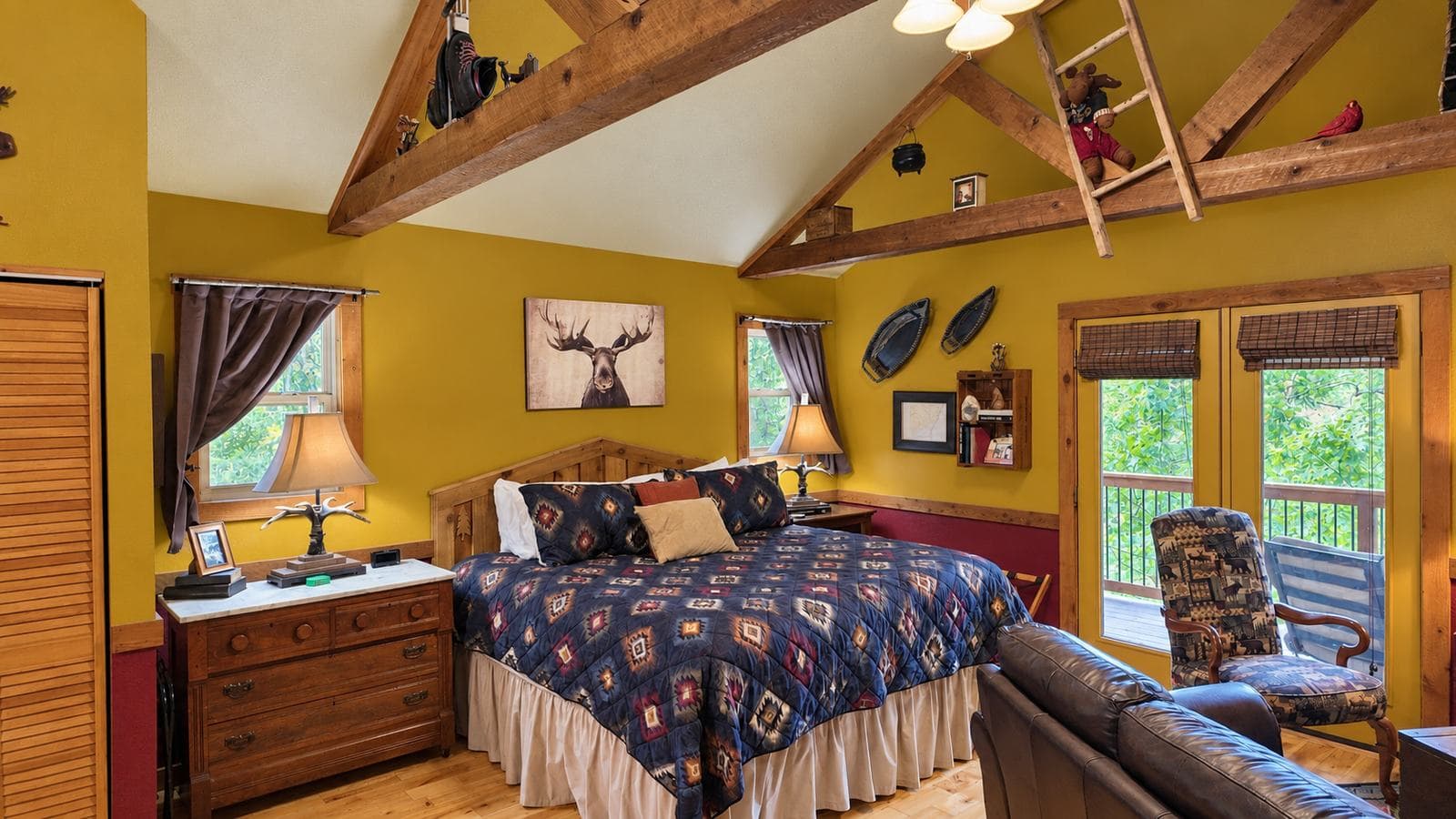 A cozy bedroom featuring a wooden bed with patterned bedding, rustic decor, and large windows overlooking greenery.