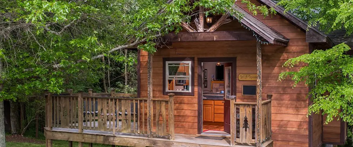 A quaint wooden cabin nestled among trees, featuring a welcoming porch and large windows.