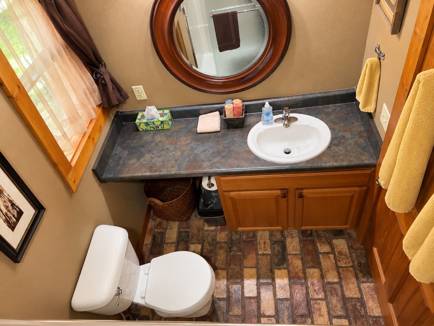 A well-lit bathroom featuring a sink with toiletries, a toilet, a round mirror, and rustic tile flooring.