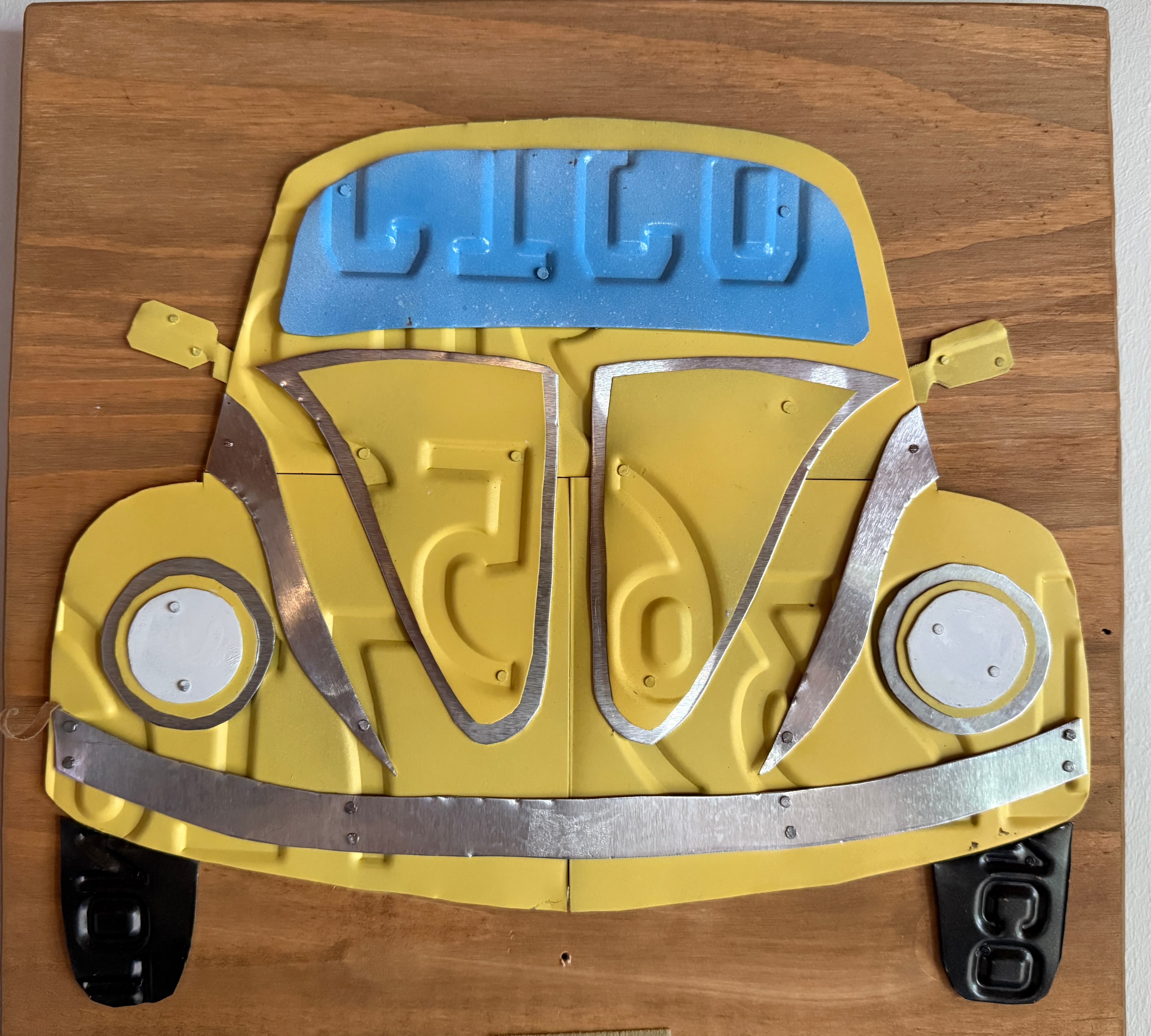 A wall-mounted art piece featuring a stylized yellow car with metal accents and a blue windshield. A wall-mounted art piece featuring a stylized yellow car with metal accents and a blue windshield.