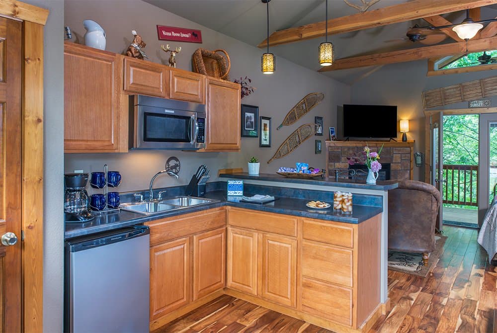 A modern kitchenette in a rustic cabin featuring light wood cabinetry, dark countertops, a stainless steel microwave, a small refrigerator, and a sink, with views into a cozy living area and an outdoor deck.