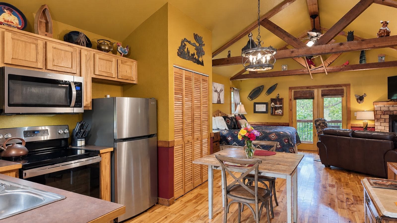 A cozy kitchen and living area with wooden beams, a dining table, and a small couch, featuring warm yellow walls and large windows.