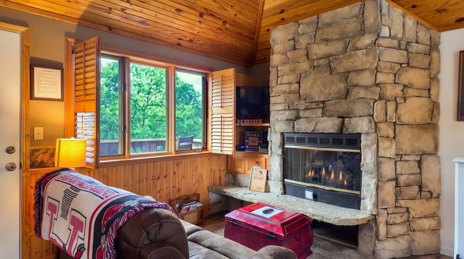 Cozy living room featuring a stone fireplace, a large window with wooden shutters, and comfortable seating.