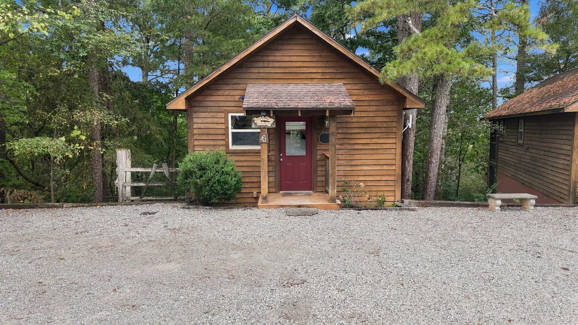 A small wooden cabin with a red door surrounded by trees and gravel.