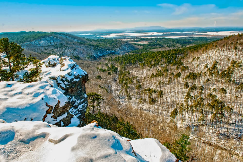 A panoramic view of a snow-covered landscape featuring hills and sparse trees under a clear blue sky.