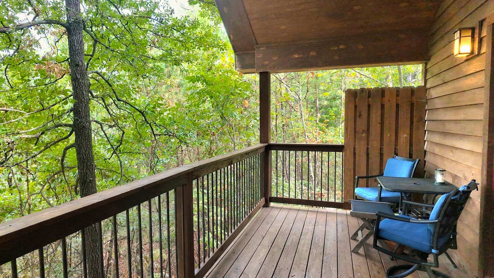 Wooden deck with two blue chairs and a small table, surrounded by lush greenery.