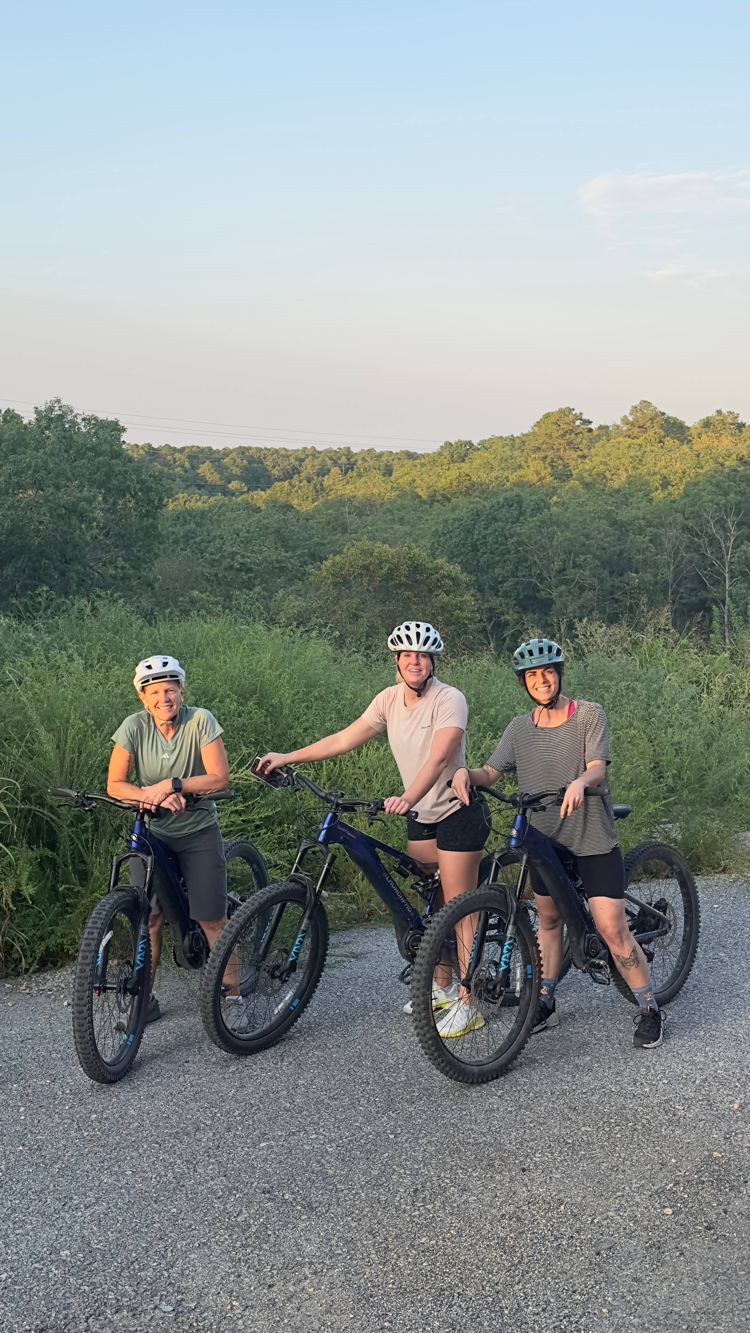 Three women pose with mountain bikes against a backdrop of trees and a clear sky.