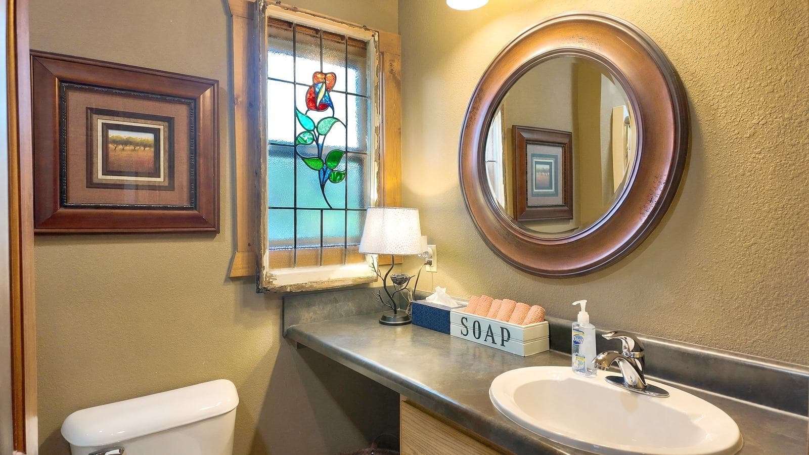 Bright bathroom featuring a stained glass window, round mirror, and decorative soap container.