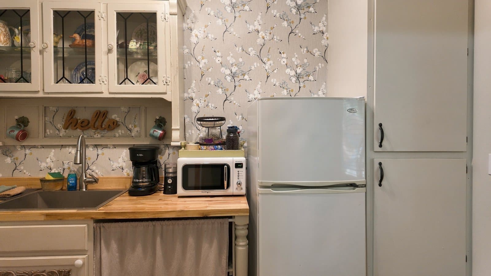 A cozy kitchen featuring a sink, coffee maker, microwave, and refrigerator against floral wallpaper.