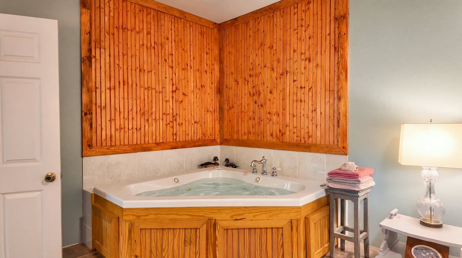 A corner jetted bathtub with wooden paneling and a lamp beside it.