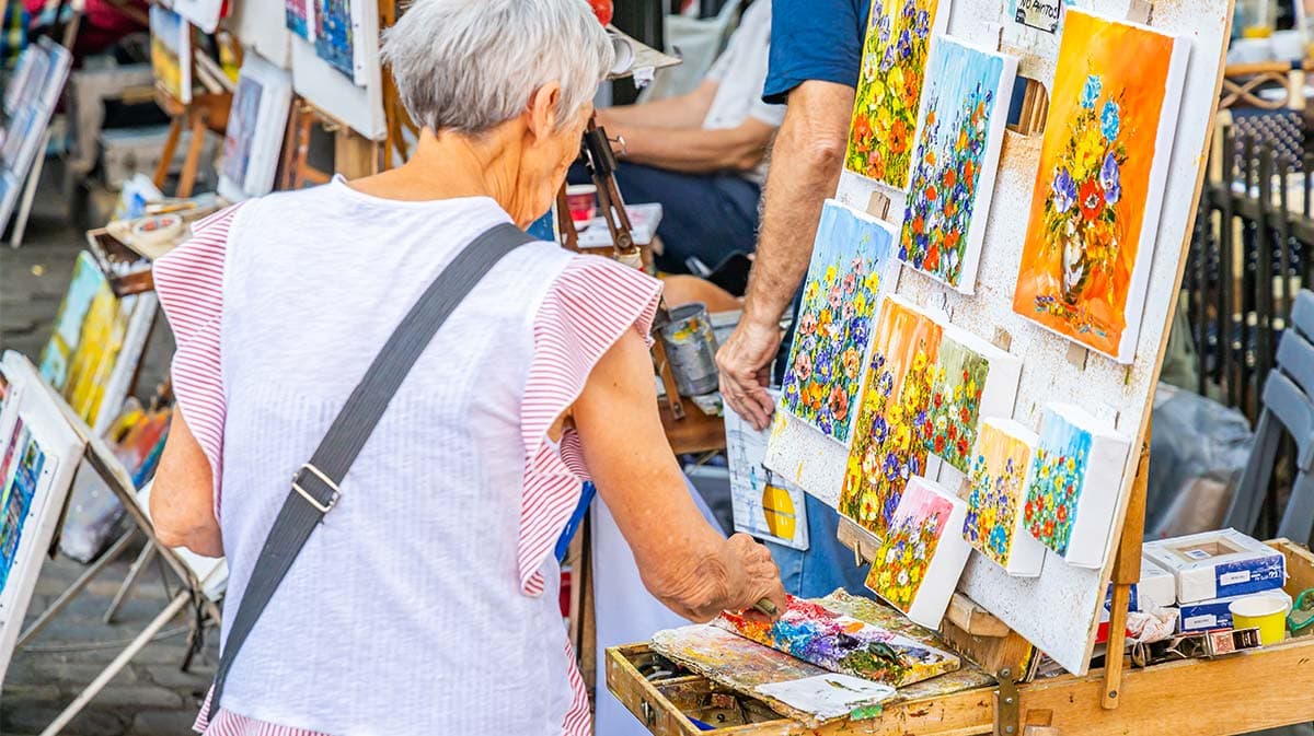 An elderly woman examines colorful paintings at an outdoor art display.
