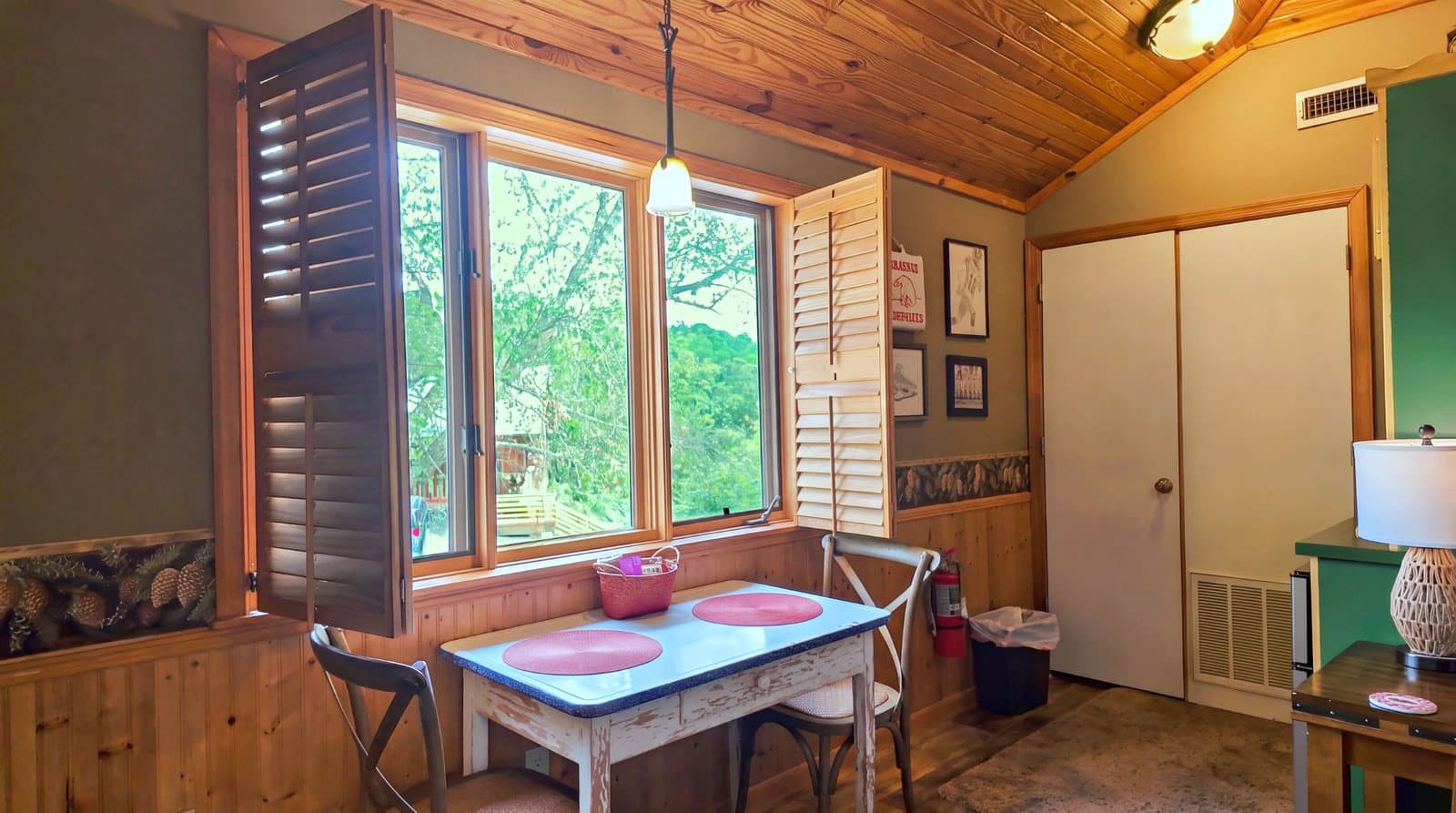 A cozy dining area with a wooden table and chairs, featuring a large window with shutters and a warm, rustic interior.
