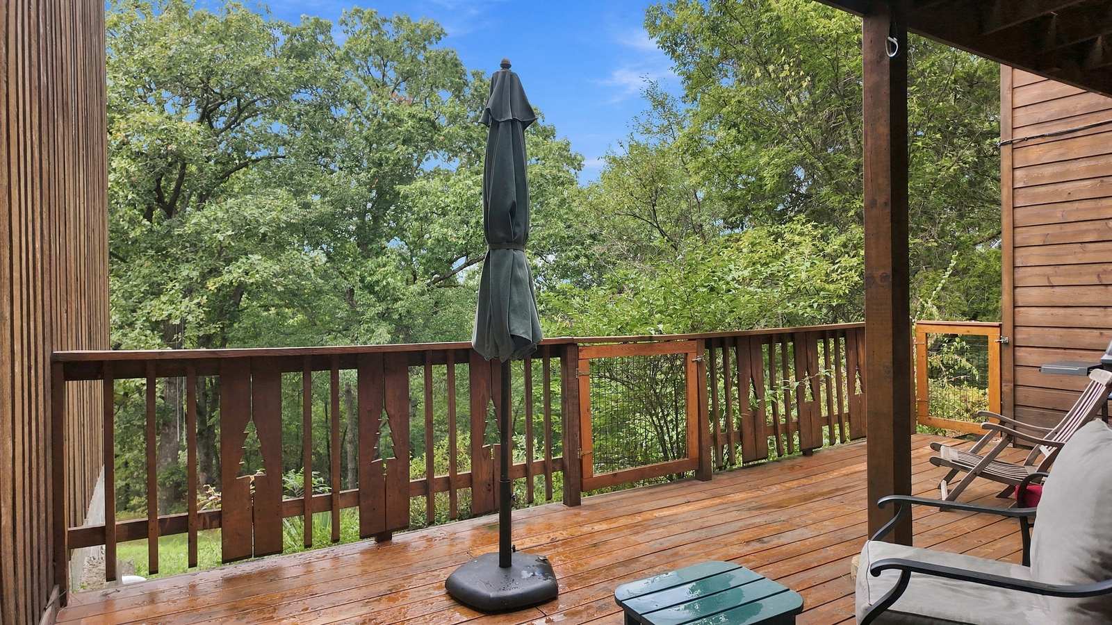 A wooden deck surrounded by green trees, featuring a tilted umbrella and a chair.