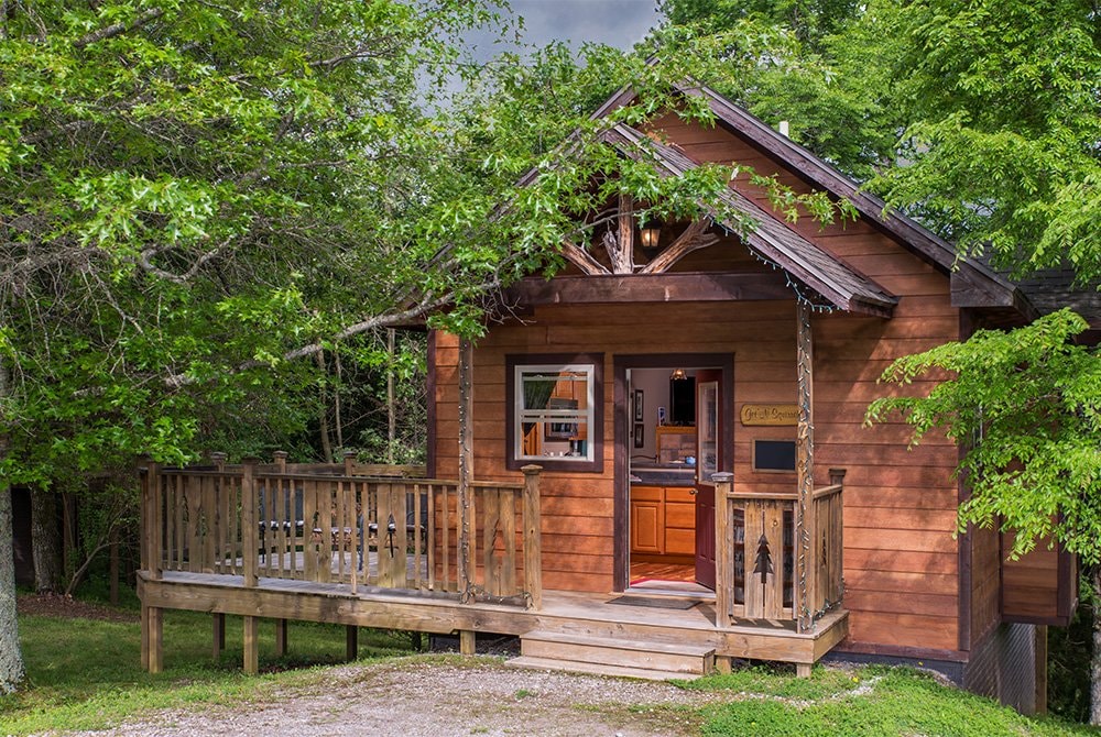 A classic wooden cabin with a gabled roof and a spacious front deck, nestled among lush green trees in a forest setting.