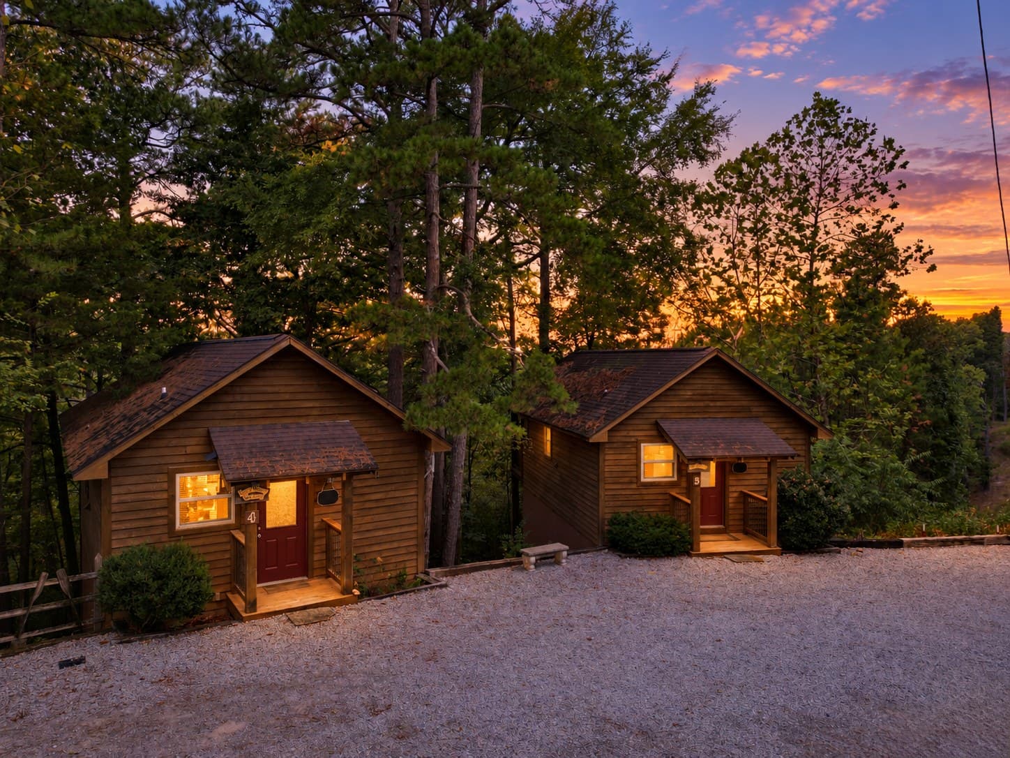 Two cozy wooden cabins illuminated by dusk, set among tall trees with a colorful sunset in the background.