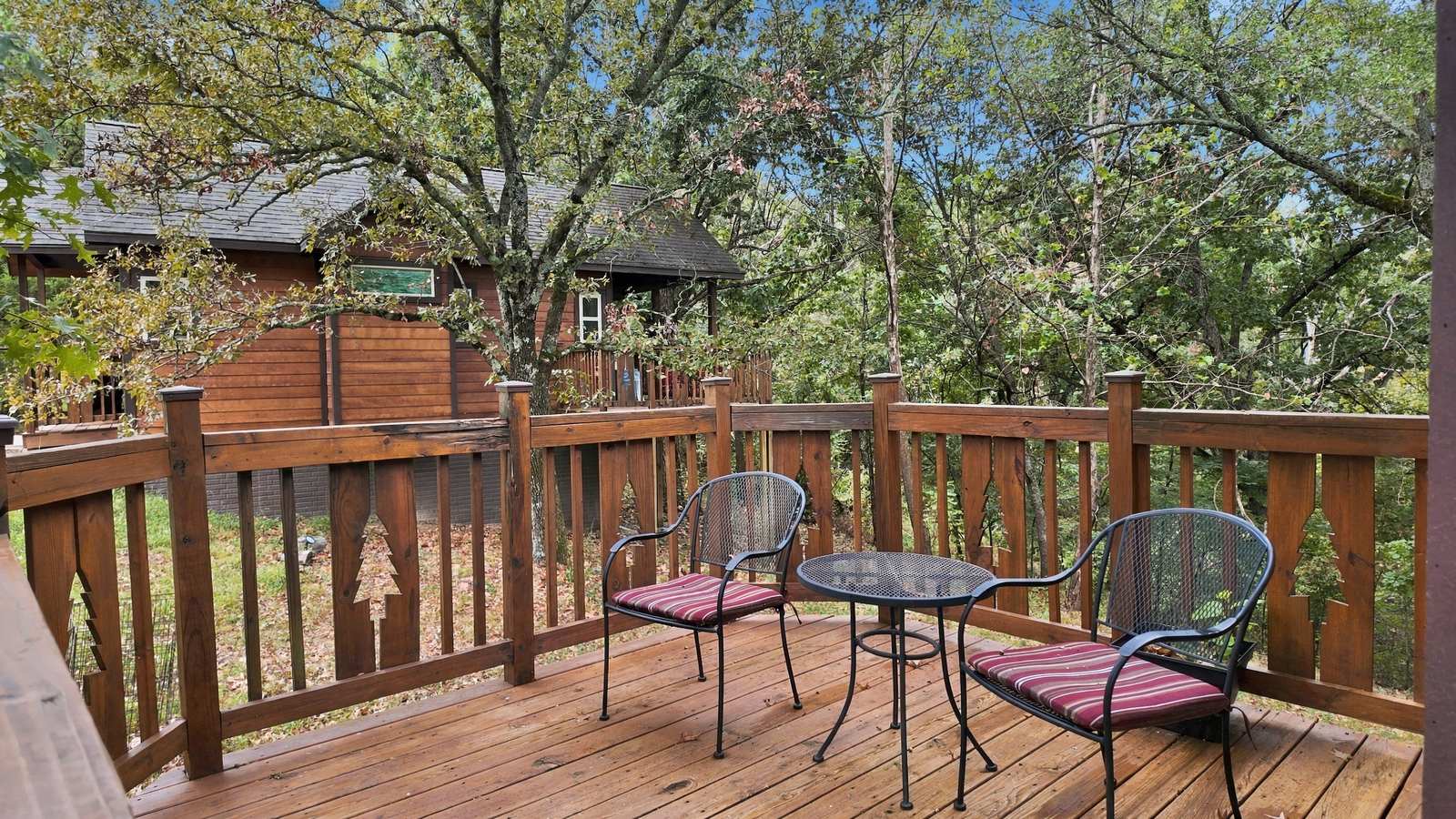 A wooden deck with two chairs and a small table surrounded by trees and a cabin in the background.